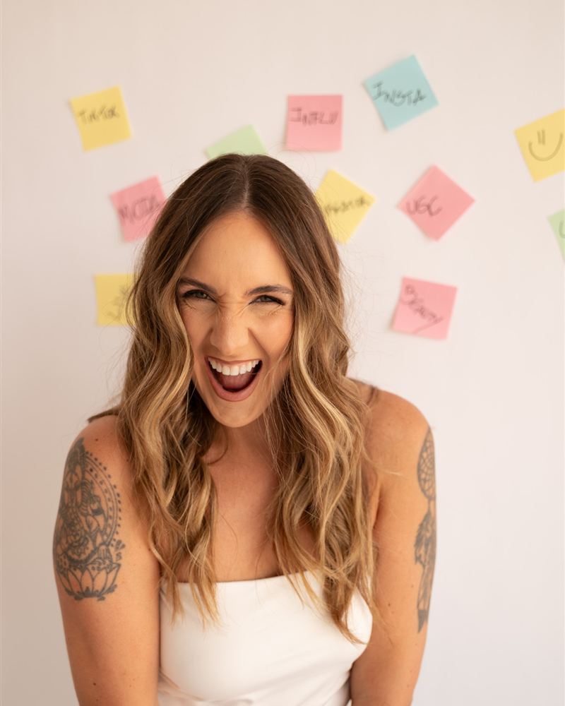 Amanda Drebes gargalhando, de blusa branca, em frente a uma parede com post-its coloridos